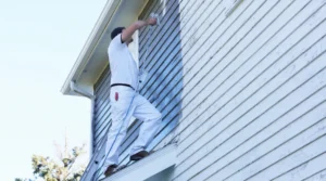 Painter standing on a ladder, applying blue paint to the exterior siding of a house with a roller.
