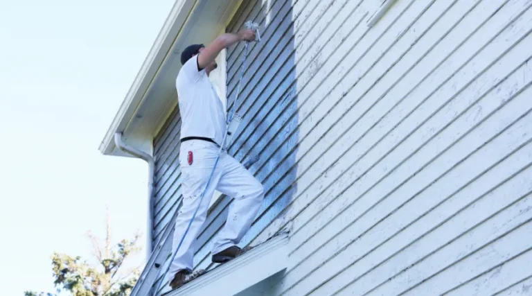 Painter standing on a ladder, applying blue paint to the exterior siding of a house with a roller.