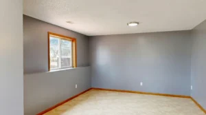 Empty gray basement room with a small window, beige tile floor, and wooden baseboards under a recessed ceiling light.
