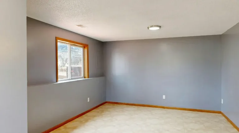 Empty gray basement room with a small window, beige tile floor, and wooden baseboards under a recessed ceiling light.
