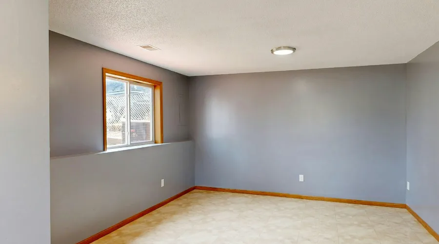 Empty gray basement room with a small window, beige tile floor, and wooden baseboards under a recessed ceiling light.