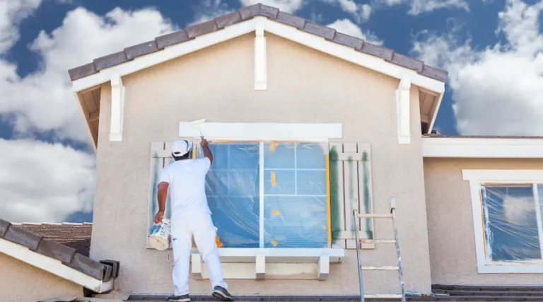 Worker in white overalls painting the window frame on a beige house, plastic sheeting protecting the window, with a ladder nearby.