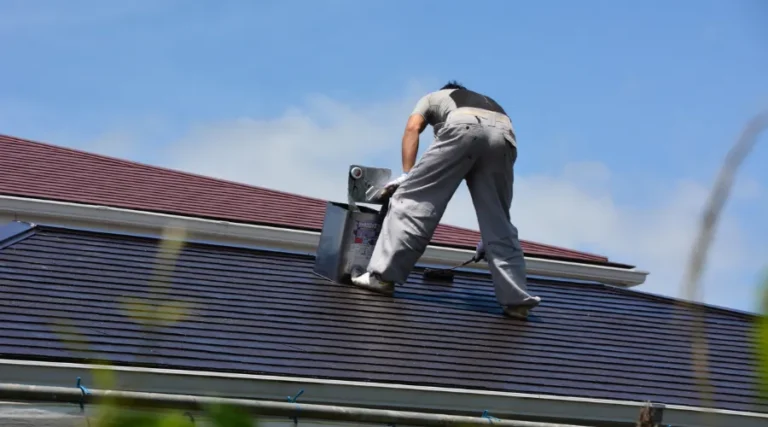 Worker on a sloped roof operating a power tool, wearing gray coveralls and a black/gray shirt from the back perspective.