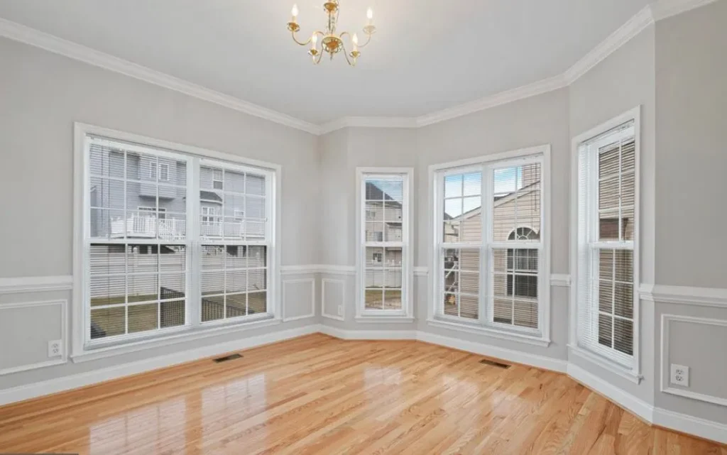Bright empty living room with light gray walls, white wainscoting, and a bay of large windows with hardwood floors.