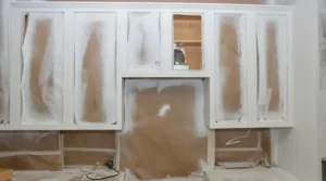White kitchen cabinets covered with masking paper for painting, with protected panels and exposed wall area in the center.