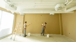 Two construction workers paint a beige wall in a bright, unfinished room; one on a ladder and the other rolling paint with a roller.
