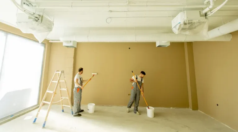 Two construction workers paint a beige wall in a bright, unfinished room; one on a ladder and the other rolling paint with a roller.