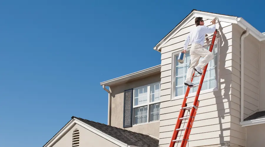 A man in a white shirt and beige pants stands on a red ladder, painting the beige siding of a two-story house against a clear blue sky near a window edge.